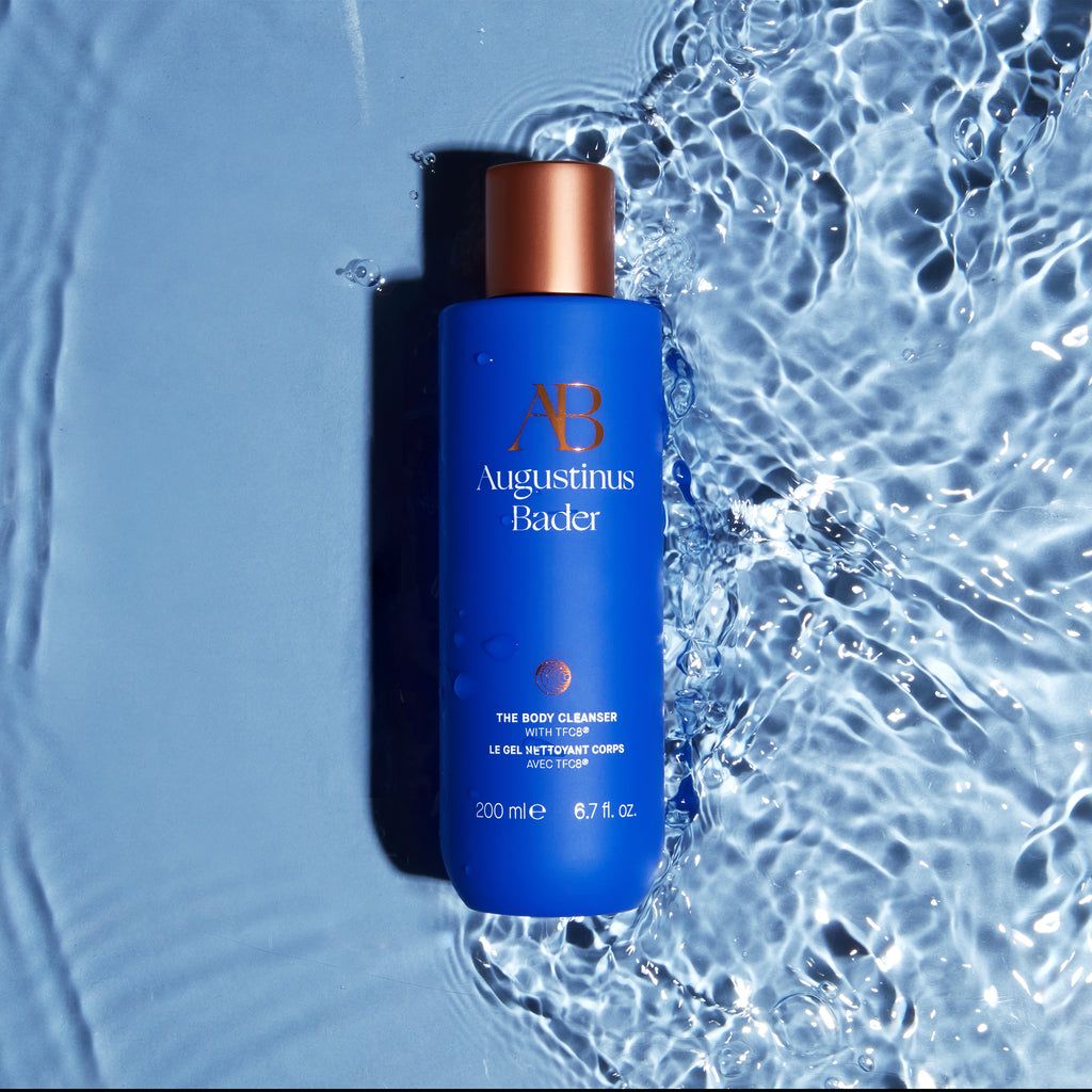 A bottle of augustinus bader body cleanser lying on a wet surface with water splashes around it.