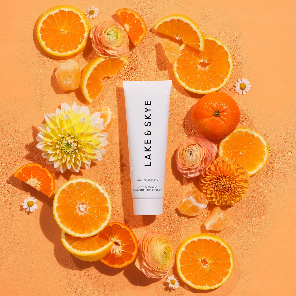 Tube of lake & skye body lotion surrounded by vibrant citrus fruits and flowers on an orange background.
