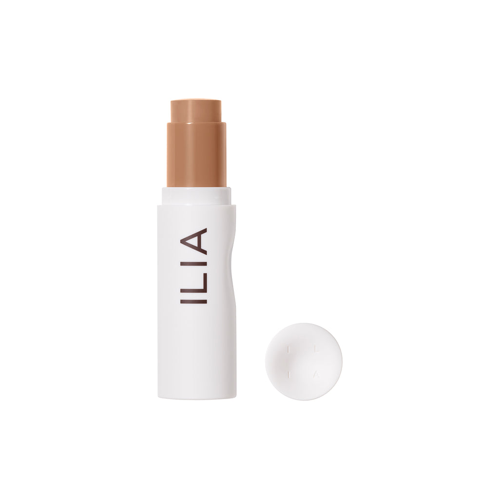 A stick of ilia brand foundation with its cap placed beside it on a white background.