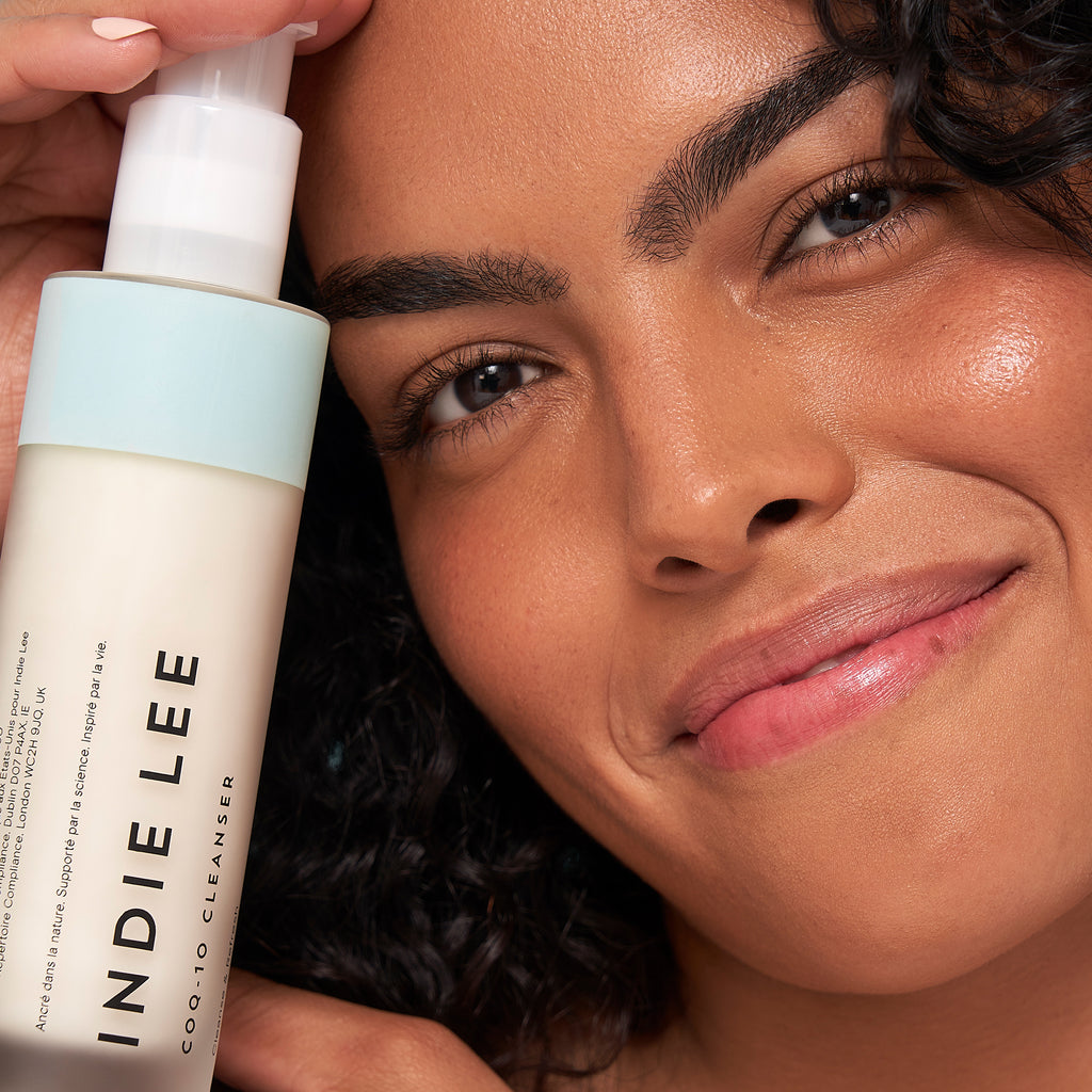 Close-up of a smiling woman holding a bottle of Indie Lee COQ-10 Cleanser near her face, highlighting the Indie Lee COQ-10 Cleanser and her clear complexion.