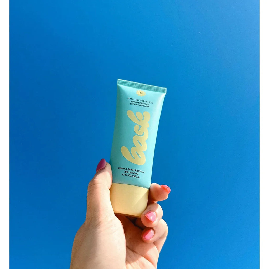 A hand holding a tube of Bask Daily Invisible Gel SPF 40 Sunscreen against a blue background.