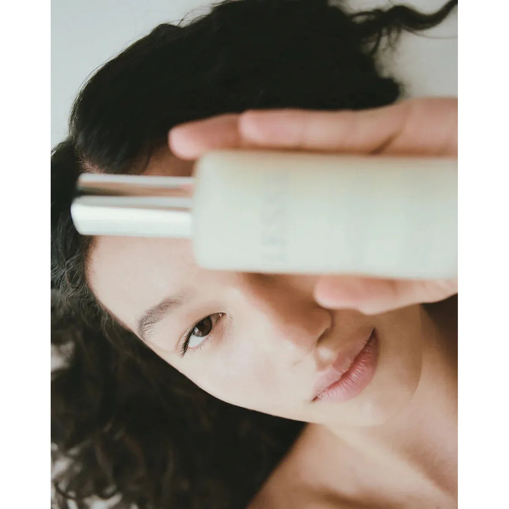 Close-up of a woman holding the LESSE Calming Cleanser near her face, looking at the camera with focused eyes, set against a white background.