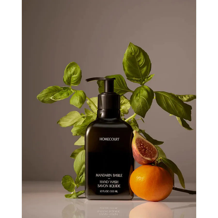 A bottle of Homecourt Hand Wash labeled "mandarin basilic" surrounded by basil leaves and a halved mandarin on a neutral background.