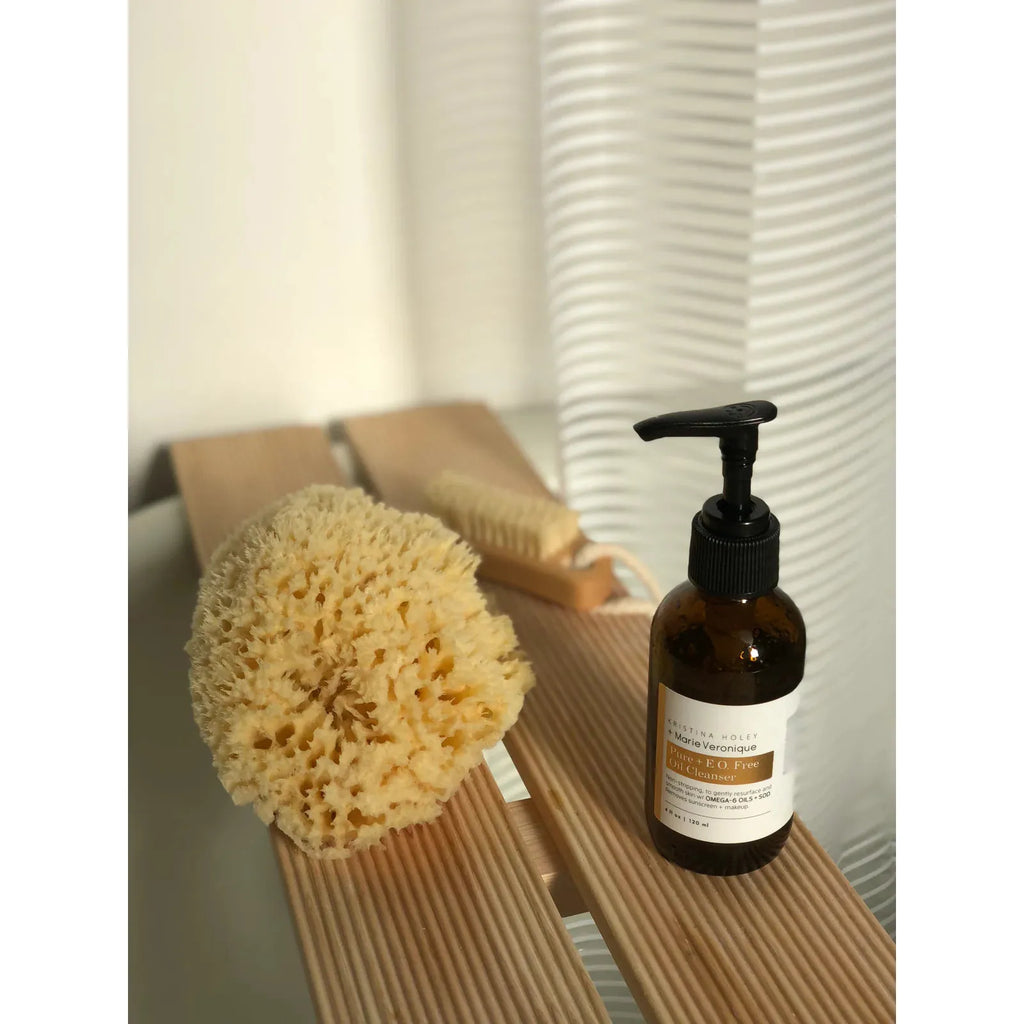 Natural sea sponge and dispenser bottle on a wooden bath tray with soft lighting and blinds in the background.