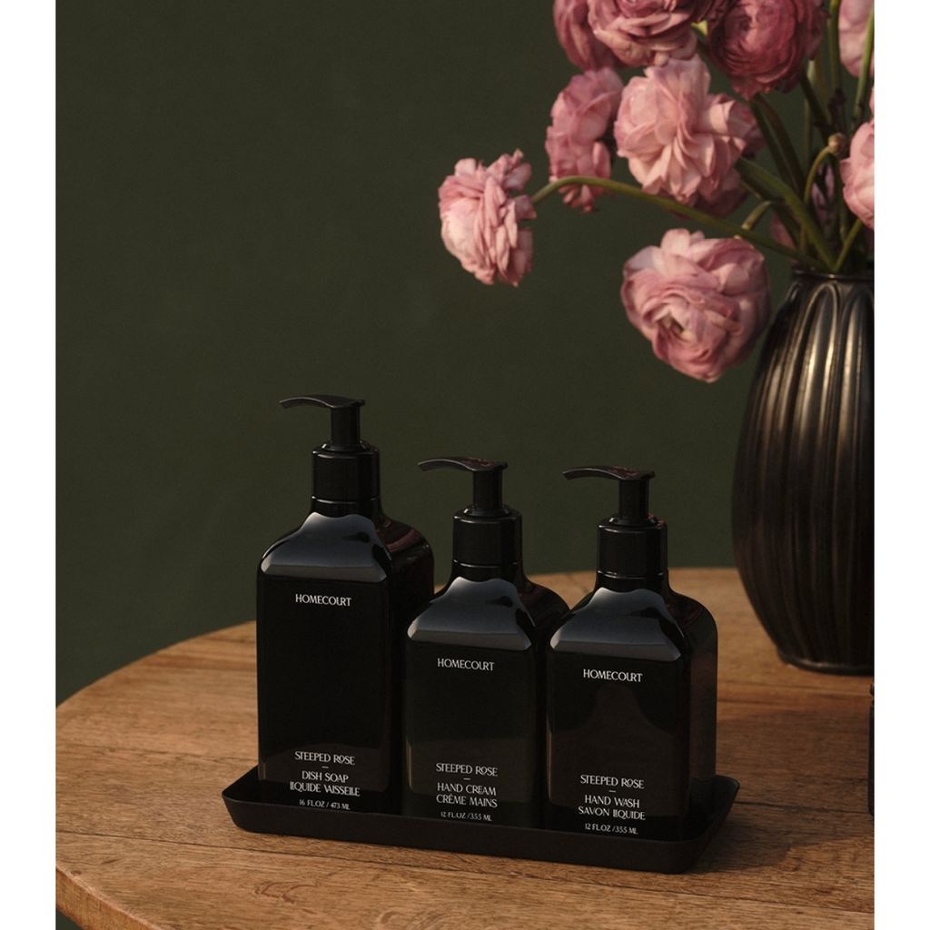 Three dark amber bottles with labels and pump dispensers on a Homecourt Yamazaki Home Steel Tray, set against a backdrop of pink flowers and a dark green wall.