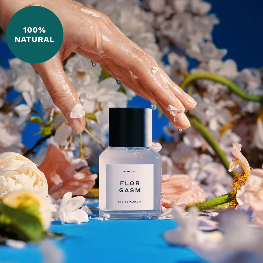 A hand with painted nails touches flowers around a perfume bottle labeled "Heretic Parfum Florgasm" against a blue backdrop, with a "100% natural jasmine fragrance" label in the corner.