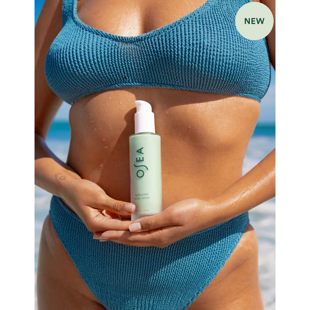 Woman in a blue bikini holding a bottle of Osea Hyaluronic Body Serum, standing against a backdrop of the sea, with a "new" label in the corner.