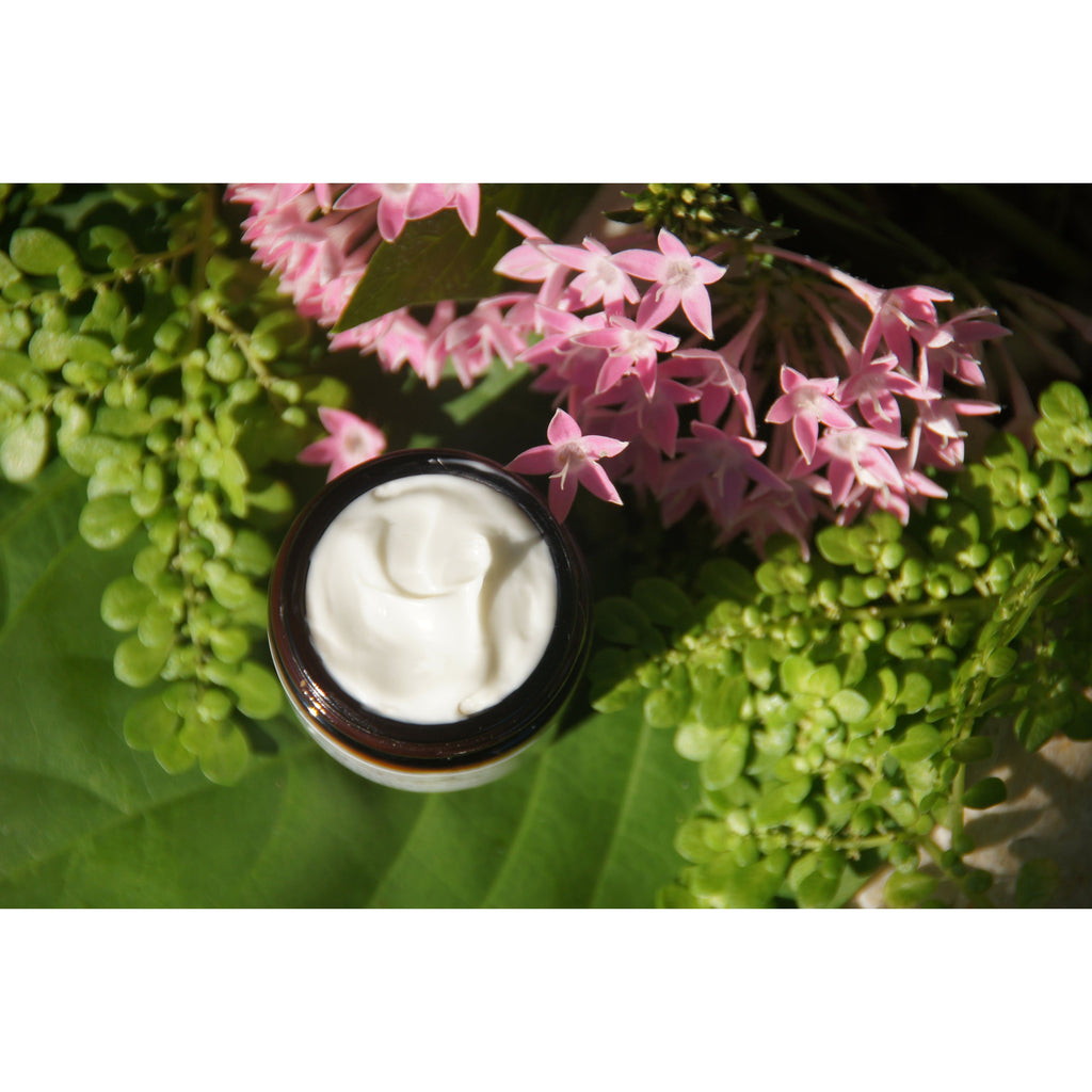 Jar of cream surrounded by pink flowers and green leaves.
