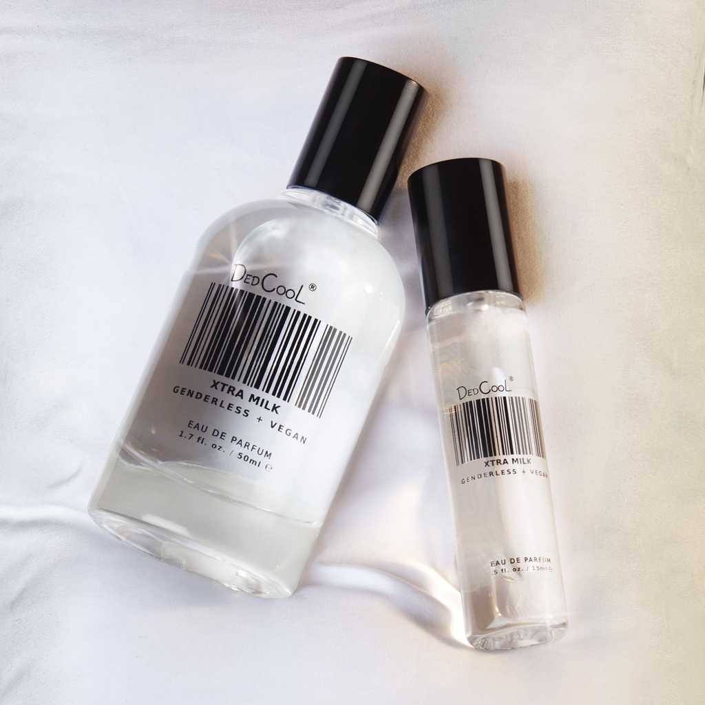 A clear bottle with a black lid of the full size DedCool Xtra Milk perfurm laying on a fluffy white background next to the clear bottle with a black lid of the travel size extra milk perfume