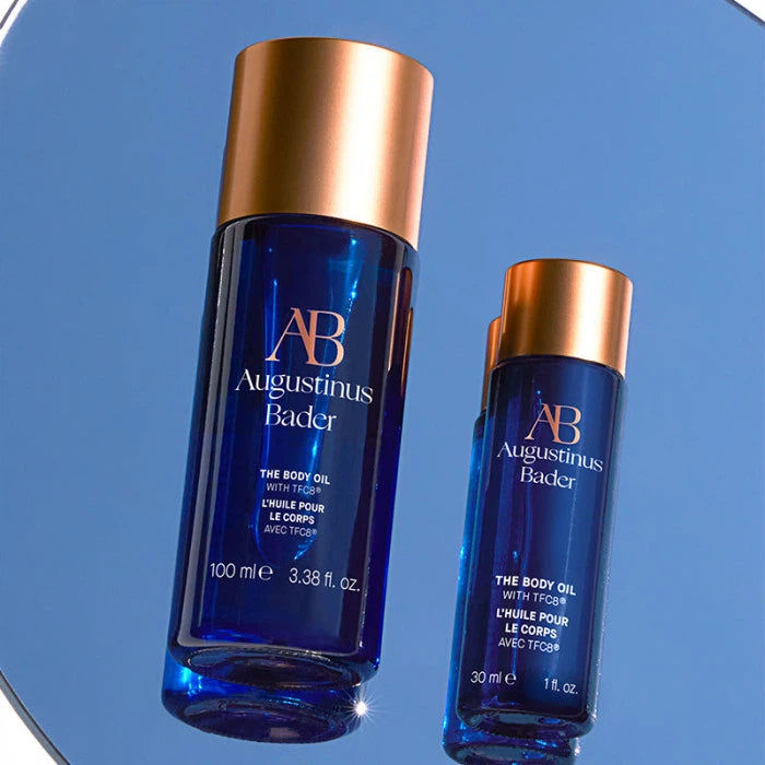 Two bottles of augustinus bader body oil in different sizes placed against a blue background.