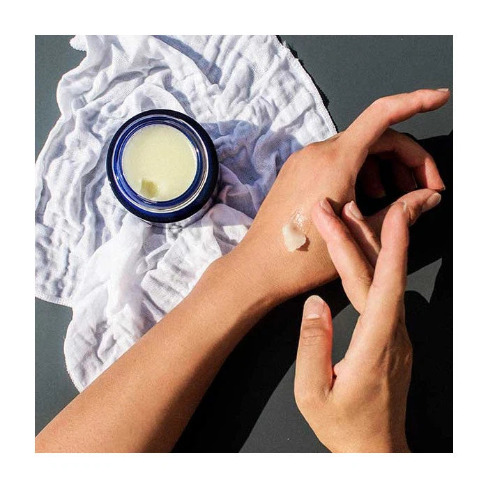 A person applying cream from a blue jar onto their hand, with a crumpled white cloth in the background.