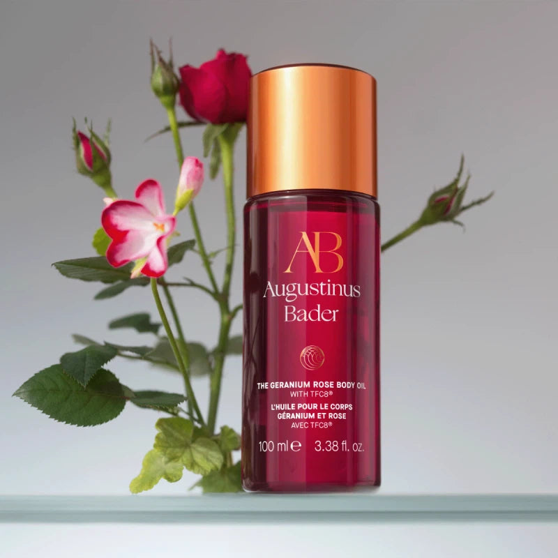 Bottle of augustinus bader geranium rose body oil with a branch of roses beside it on a neutral background.