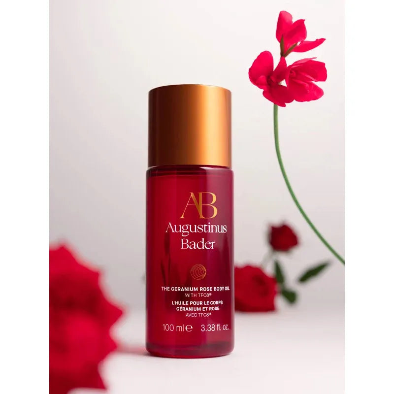 A bottle of augustinus bader the geranium rose body oil with a pink flower in the background.