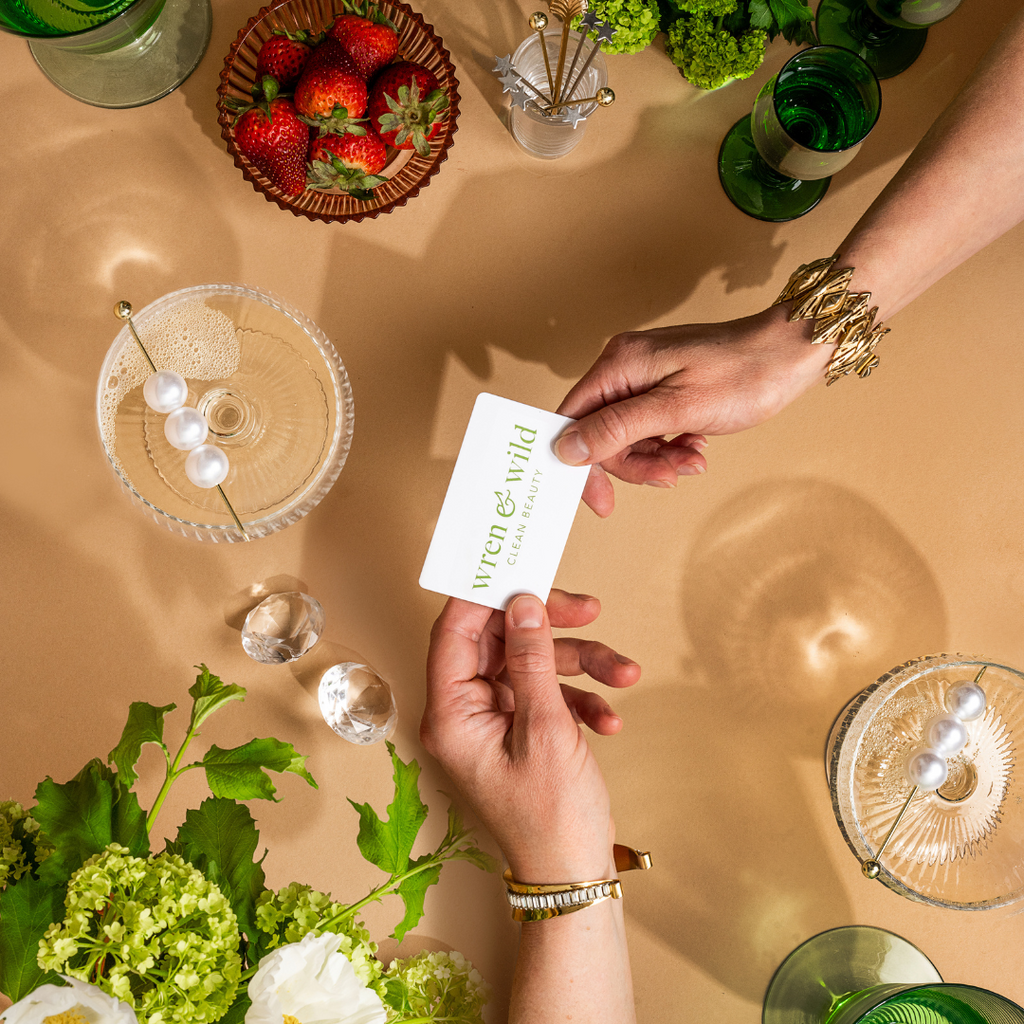 Two hands exchanging a Wren and Wild Gift Card over a table adorned with drinks, strawberries, and decorative items on a beige background.