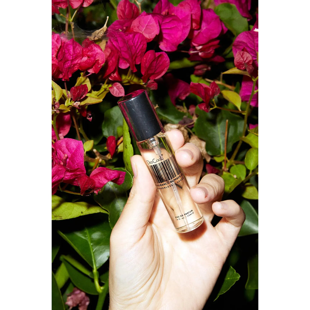 A hand holding a tube of concealer in front of a bush with pink flowers.