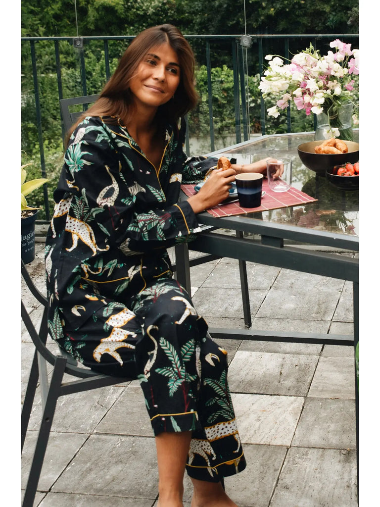 A person in Germaine des Prés Camille Black Jungle Print Pajamas sits on a patio chair, holding a cup, with a table set for breakfast including flowers, fruit, and bread.