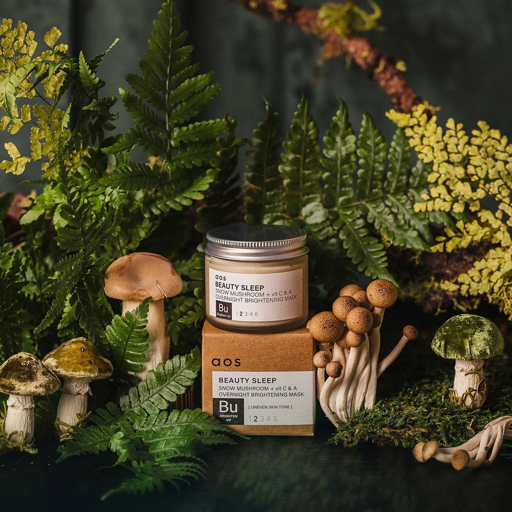 A skincare product surrounded by mushrooms and foliage on a dark background.