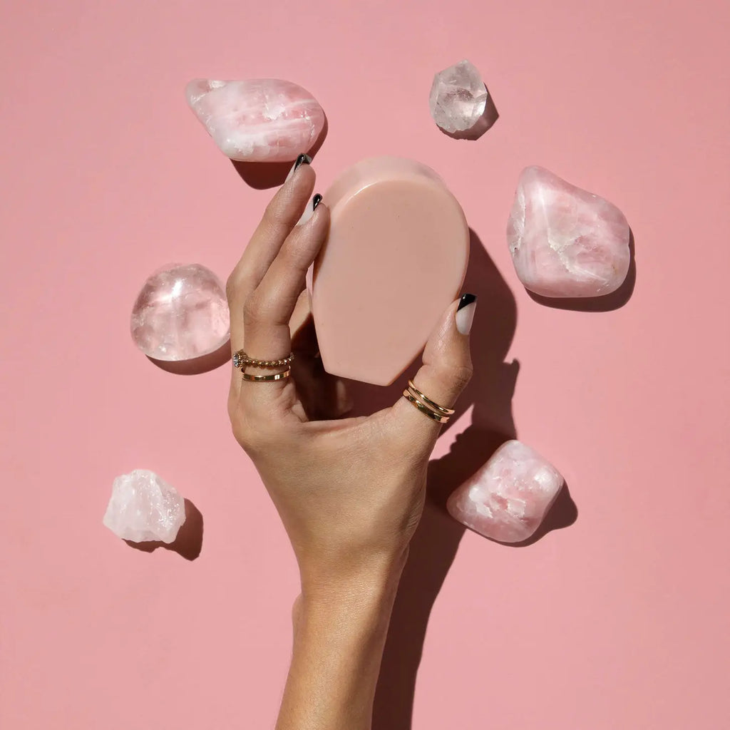 A hand with polished nails surrounded by rose quartz crystals against a pink background.