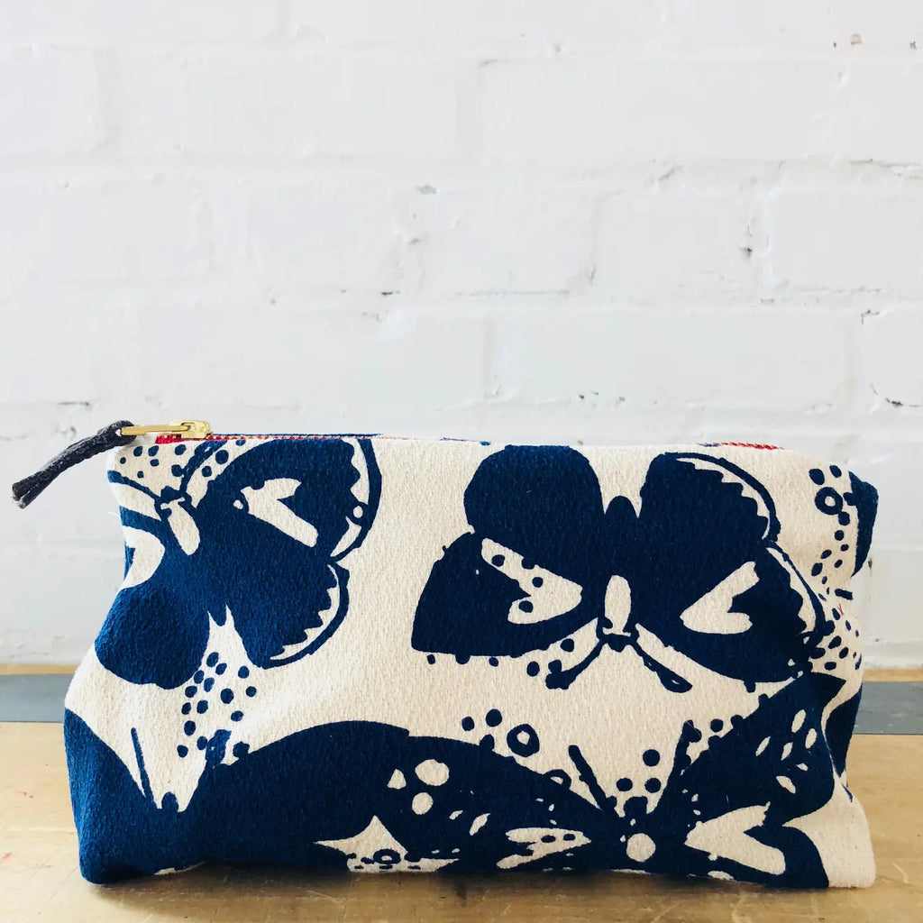 A white cosmetic bag with a blue floral pattern placed in front of a white brick wall.