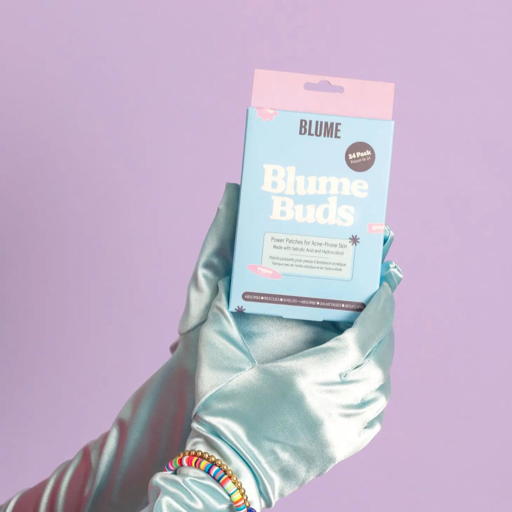 A hand wearing a teal satin glove holds a box of blume buds acne patches against a purple background.