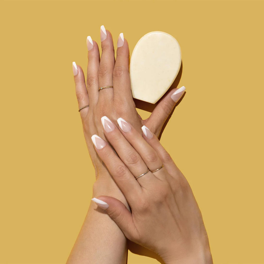 Two hands with pink manicured nails holding a bar of soap against a plain yellow background.