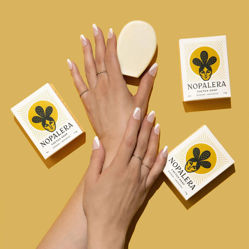 A person's elegantly manicured hands presenting a bar of soap with its packaging on a yellow background.