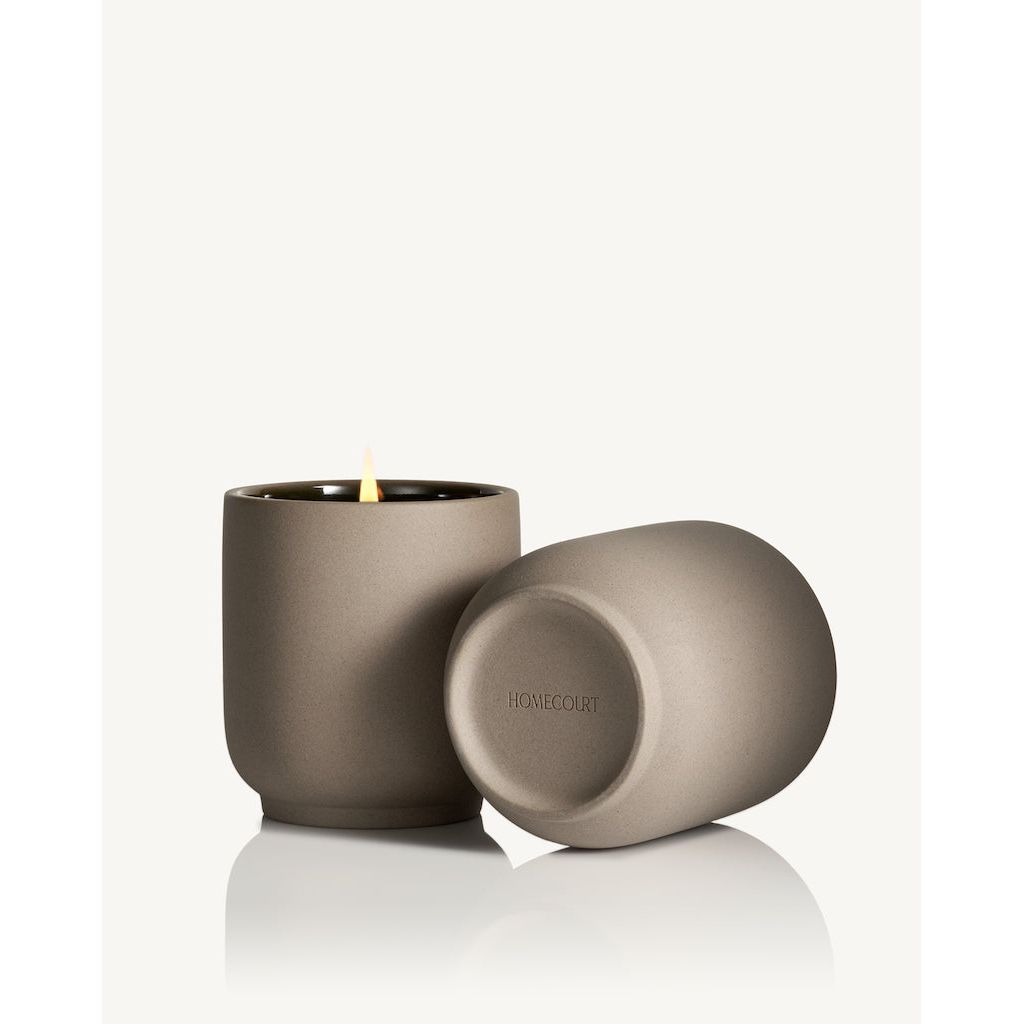 A lit candle in a matte grey holder with a matching lid placed beside it.
