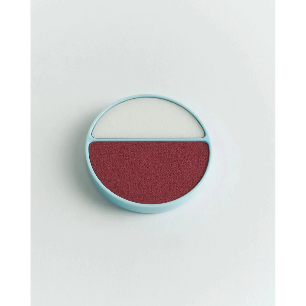 A round, dual-color cosmetic blush palate with one half white and the other half burgundy, against a pale background.