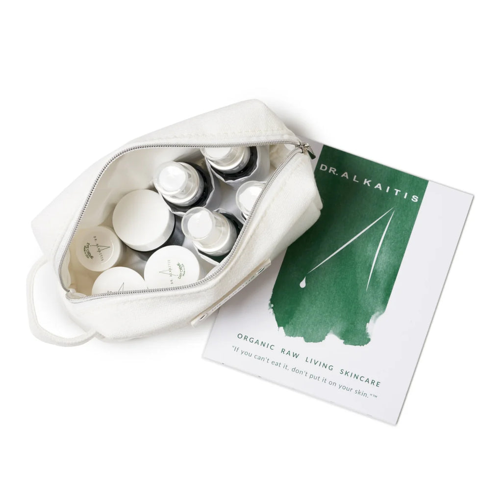 A white toiletry bag with various skincare product containers, perfect for eco-conscious travelers, next to a brochure titled "Dr. Alkaitis Organic Raw Living Skincare" with a green and white cover. This Dr. Alkaitis Organic Travel Kit Deluxe is convenient and TSA-approved for smooth, eco-friendly journeys.