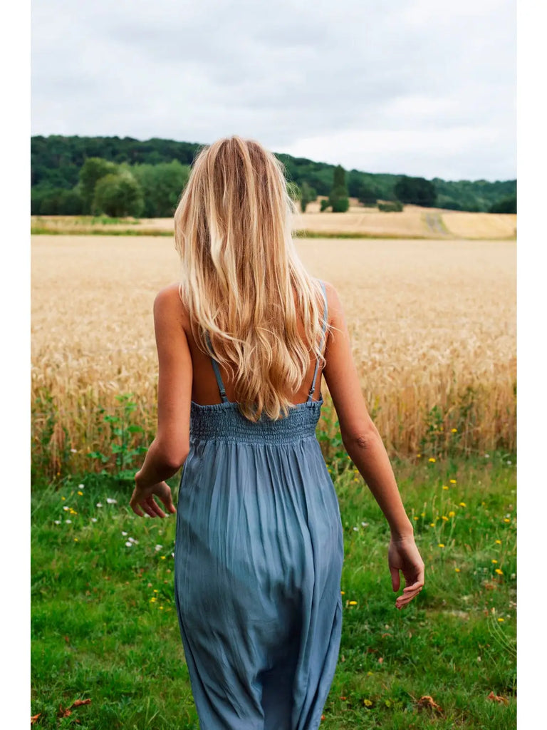 A person with long blond hair wearing the elegant Germaine des Prés Josephine Grey Nightie walks towards a field of wheat on a cloudy day.