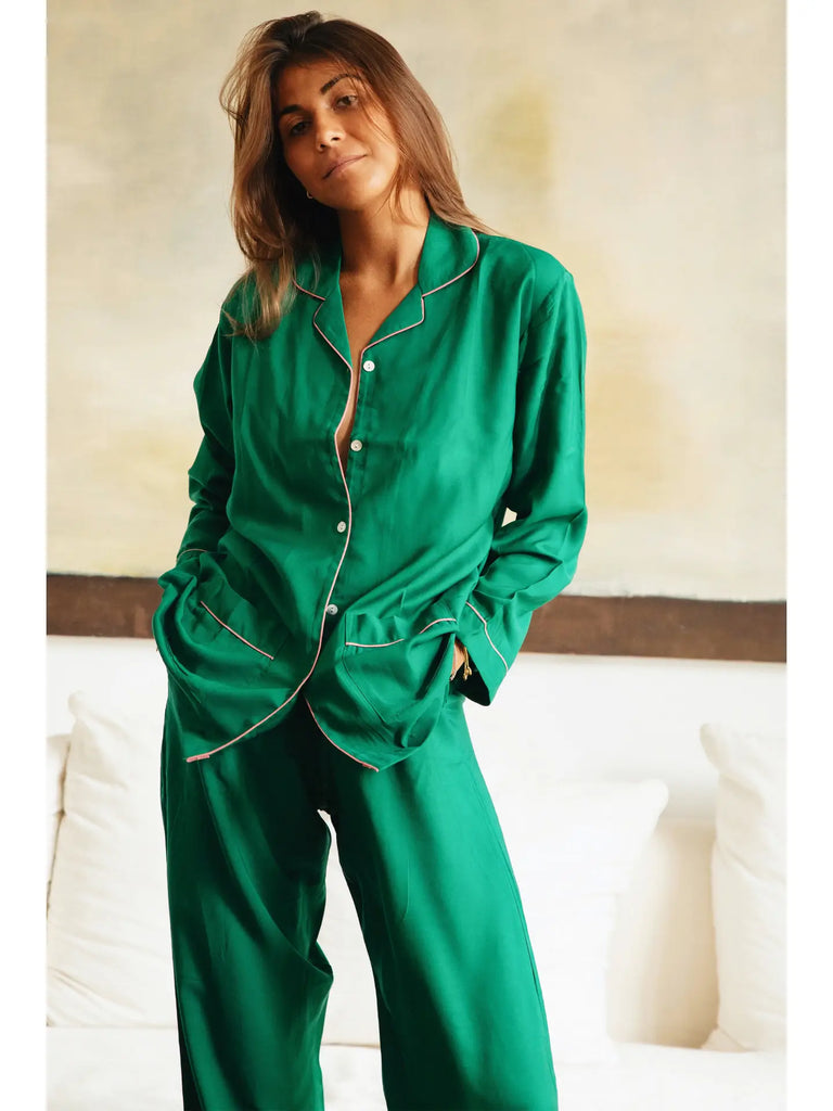 A person stands indoors, facing the camera, wearing the Germaine des Prés Camille Emerald Pyjamas with their hands in the pockets. The classic shape of the cotton and silk pajamas adds a touch of elegance to their casual look.