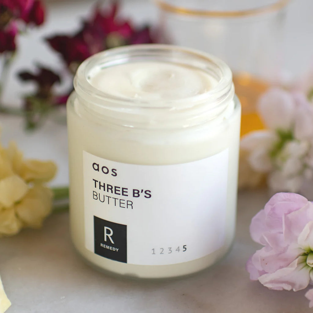 Open jar of cosmetic cream with label, surrounded by flowers.