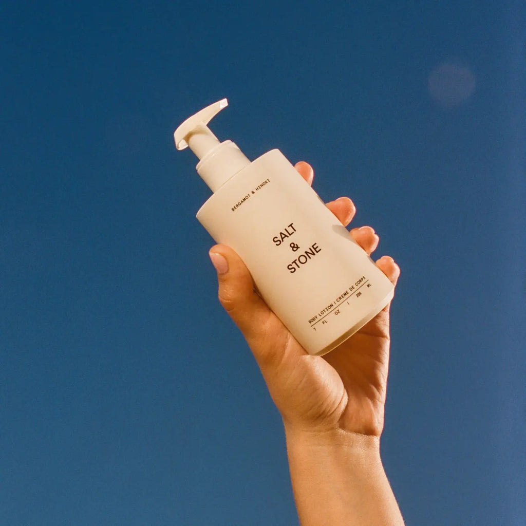 A hand holding a bottle of salt & stone sunscreen against a clear blue sky.