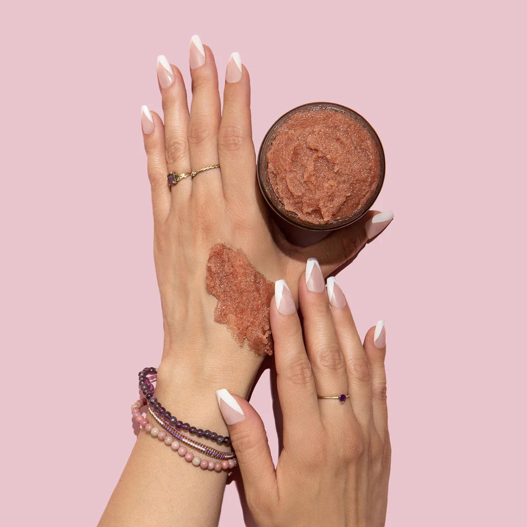 A person demonstrating a beauty scrub on their hand against a pink background.