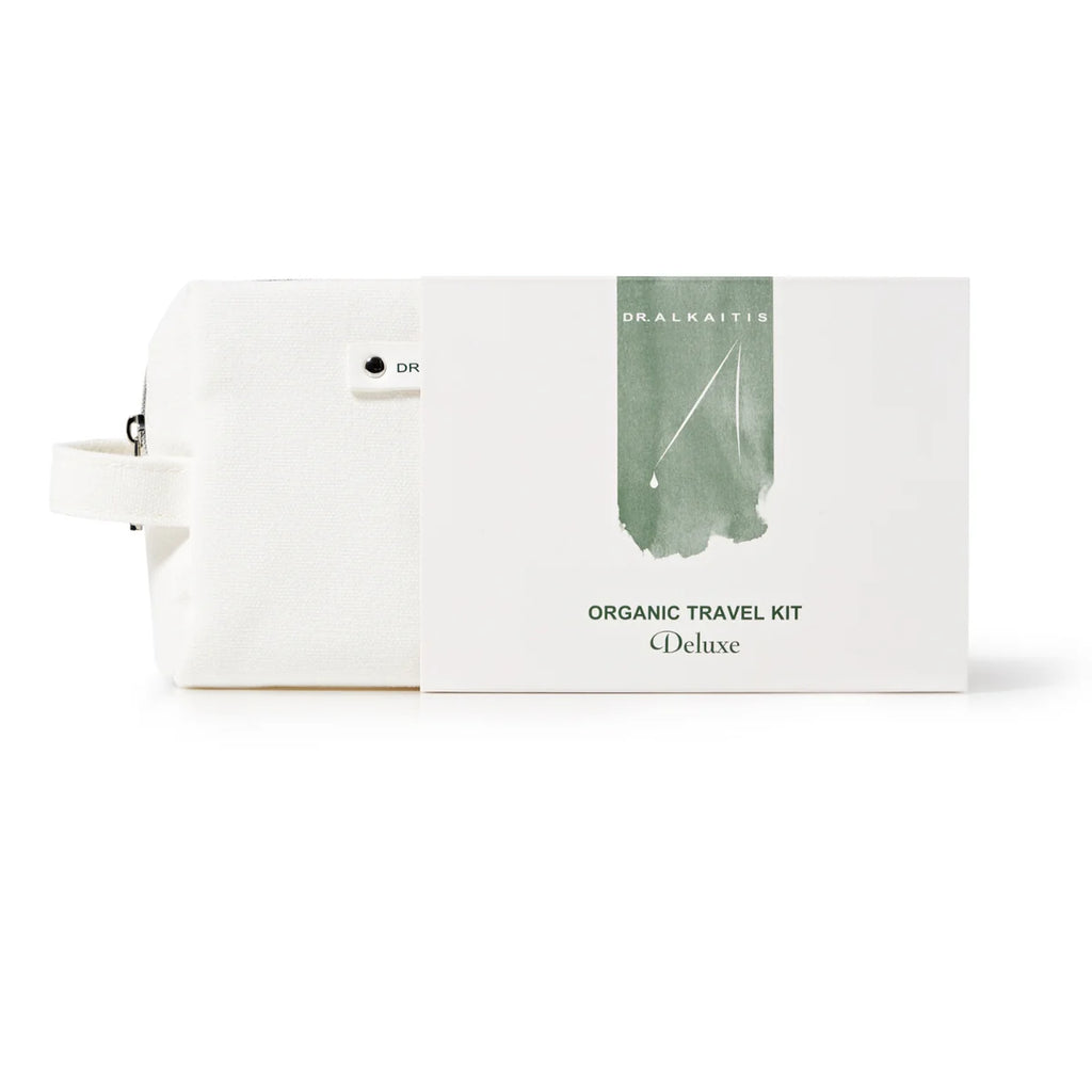 A white toiletry bag is displayed next to a white card that reads "Dr. Alkaitis Organic Travel Kit Deluxe" with green brushstroke and small text, "Dr. Alkaitis." Perfect for eco-conscious travelers, this TSA-approved kit combines style and sustainability.