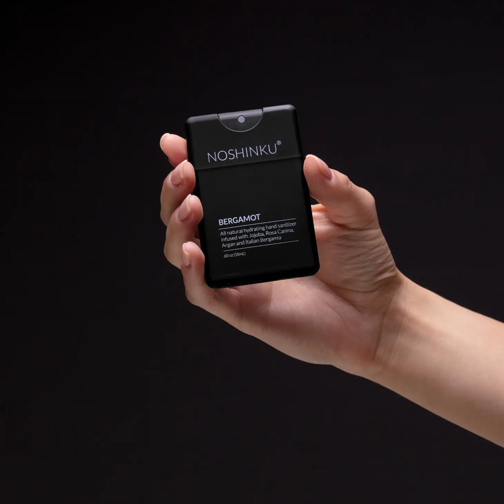 A hand holding a noshinku bergamot hand sanitizer spray bottle against a dark background.