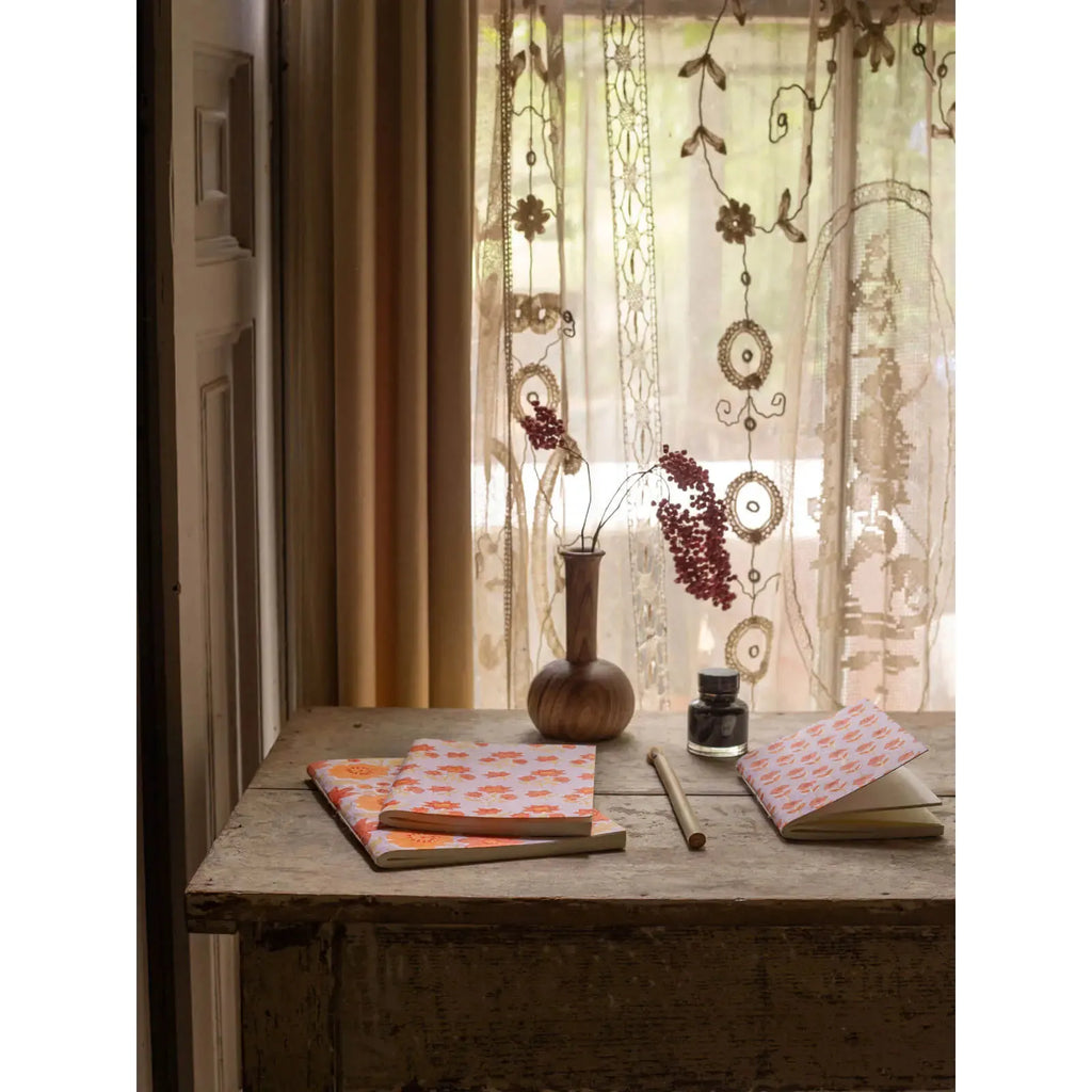 A cozy writing nook featuring a wooden desk with a vase of dried flowers, a notebook, pen, and ink by a window with lace curtains.