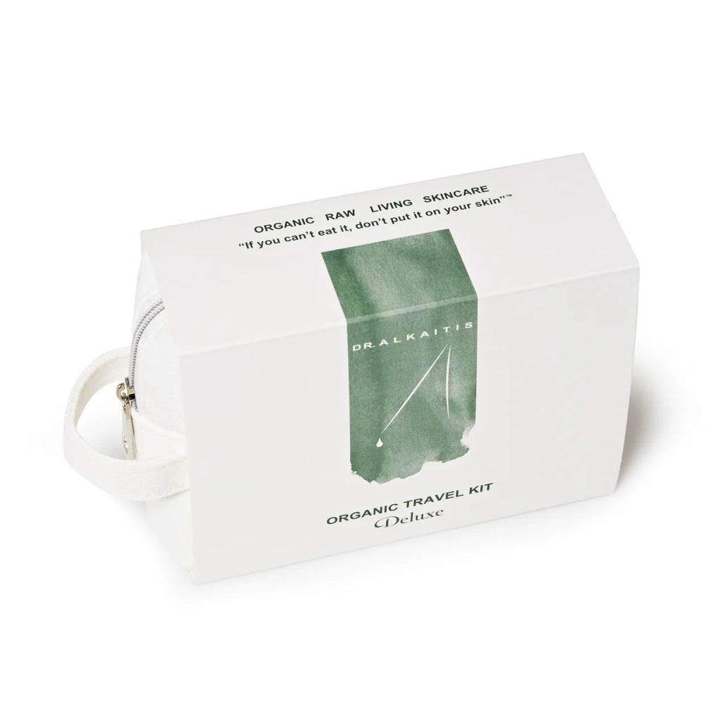 A white rectangular box with green text reading "Dr. Alkaitis Organic Travel Kit Deluxe," designed for eco-conscious travelers. Inside, a white zippered pouch holds TSA-approved organic, raw, living skincare products.