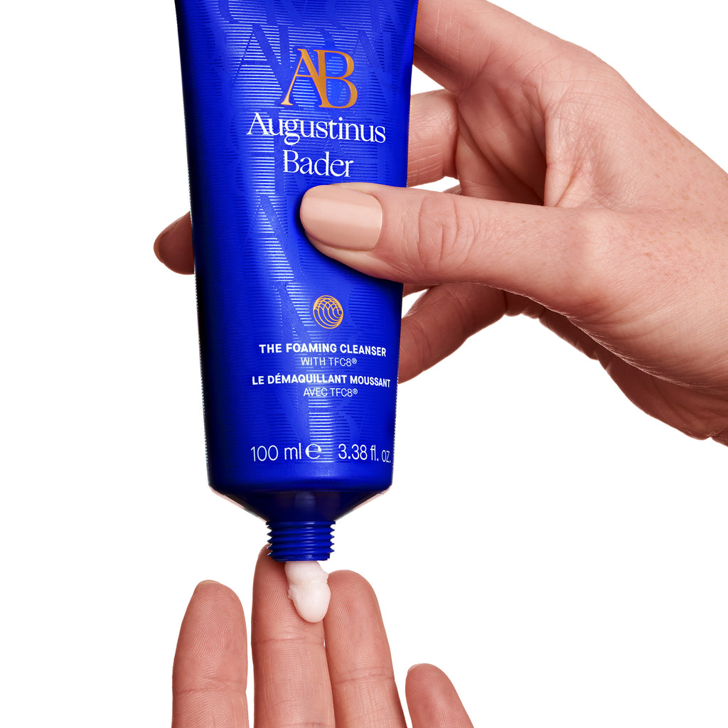 A hand squeezing a tube of augustinus bader facial cleanser against a white background.