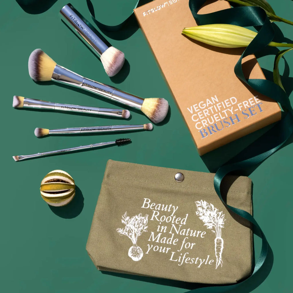 Introducing the Fitglow Beauty Master Brush Set: a vegan certified, cruelty-free collection featuring six essential makeup brushes and an eco makeup bag. The set is elegantly presented with a green pouch, citrus fruit, a beautiful flower, and a product box on a green background adorned with satin ribbon.