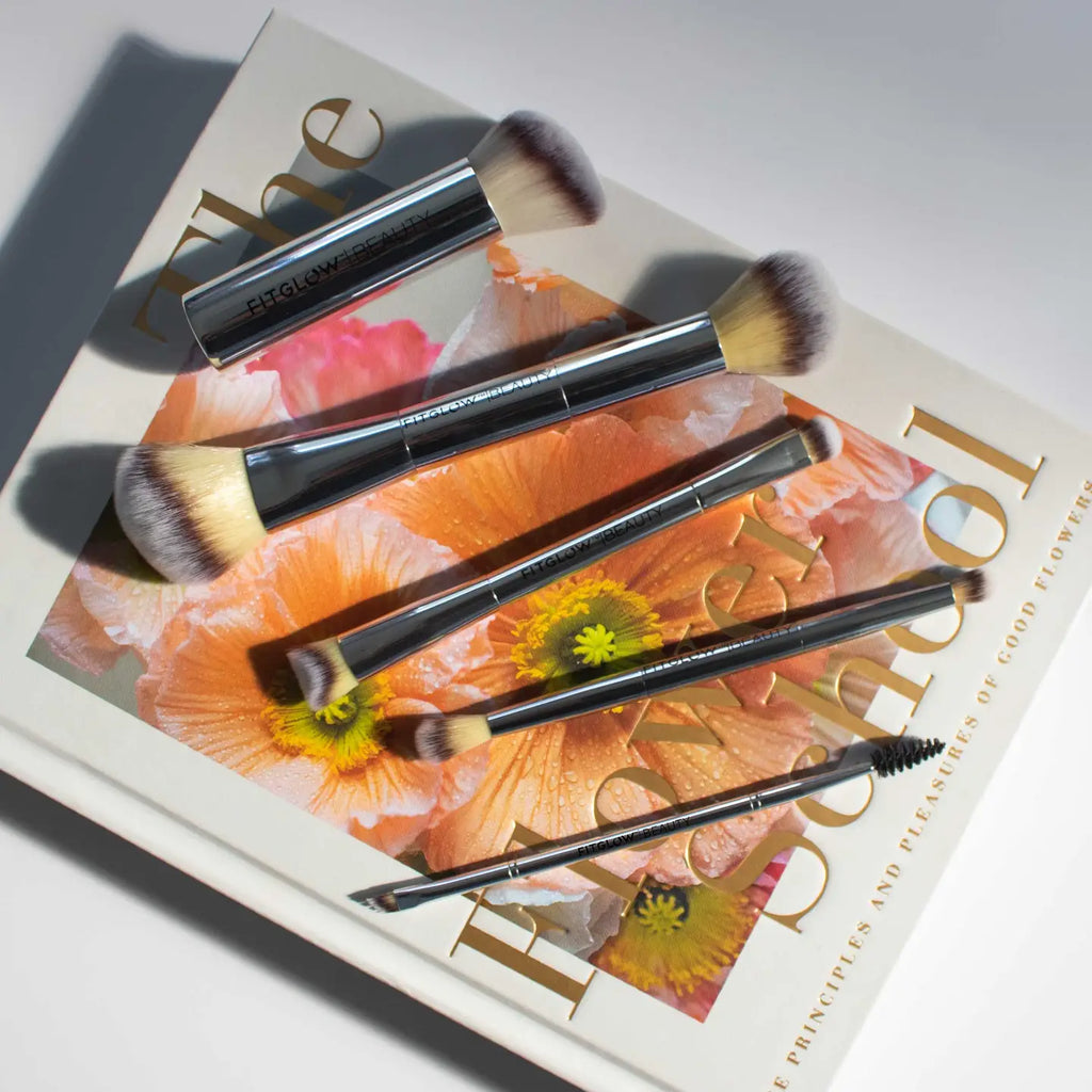 The Fitglow Beauty Master Brush Set, featuring cruelty-free brushes with metallic handles, is placed on an open book displaying vibrant flower illustrations.