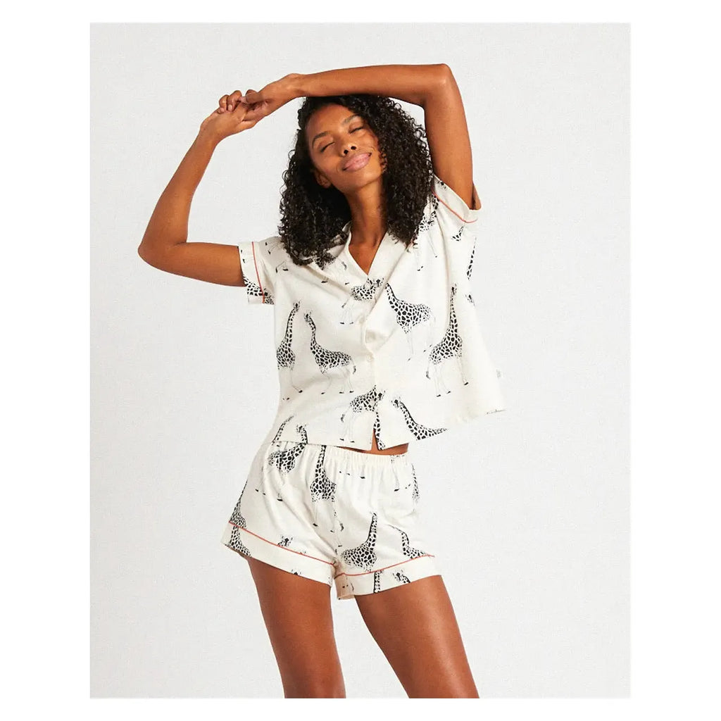 A person with curly hair stretches, wearing a white pajama set with giraffe prints. The Chelsea Peers Organic Cotton Giraffe Print includes a short-sleeve top and shorts.