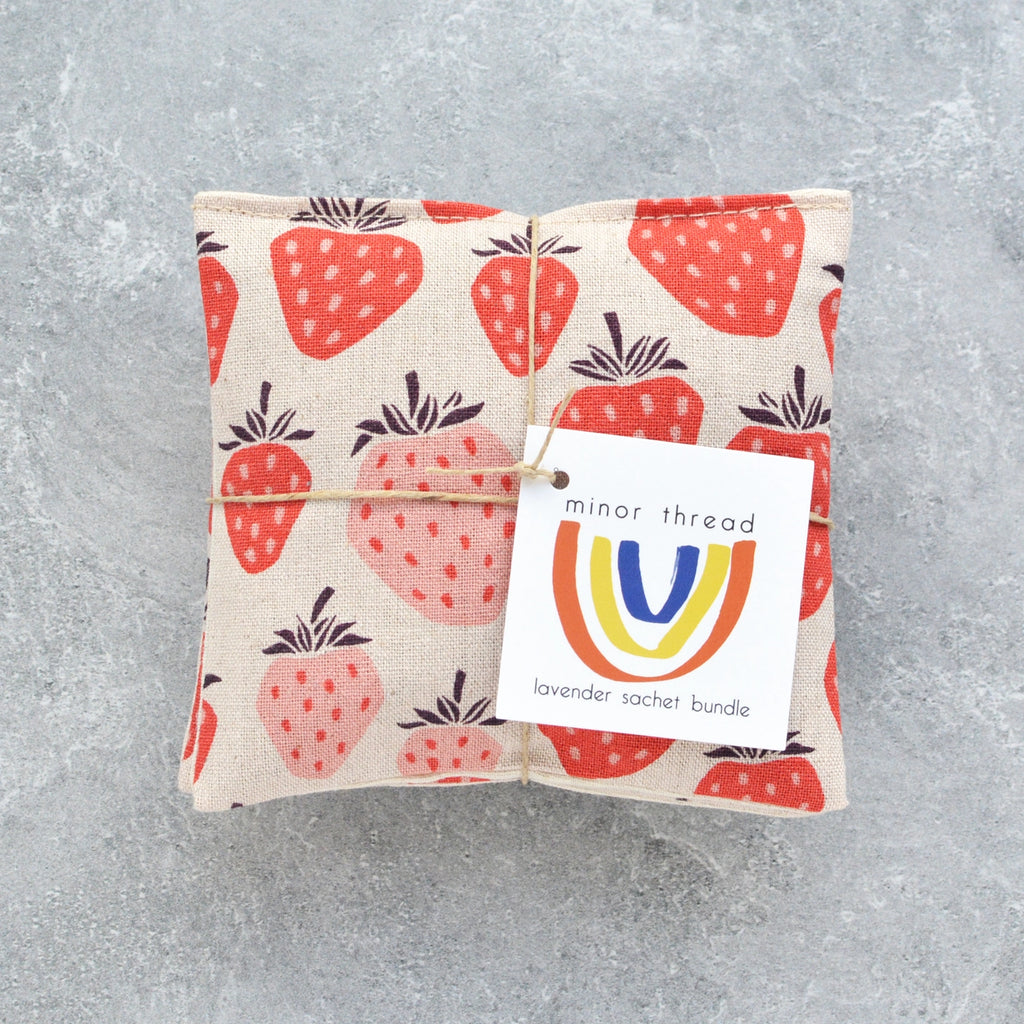 A fabric lavender sachet with strawberry print and a branded label on a textured gray background.