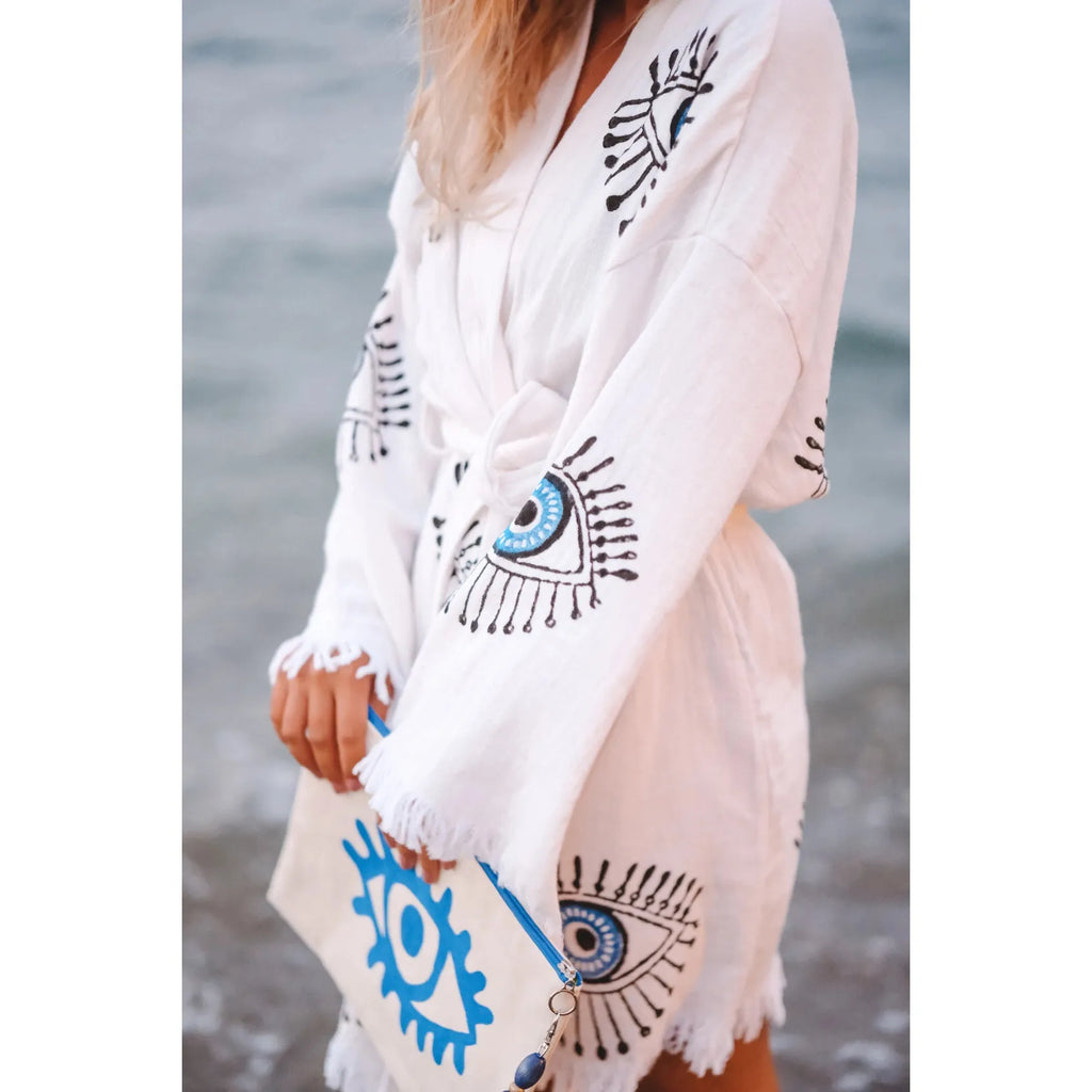 A person wearing a white bohemian-style dress with eye patterns near a body of water.