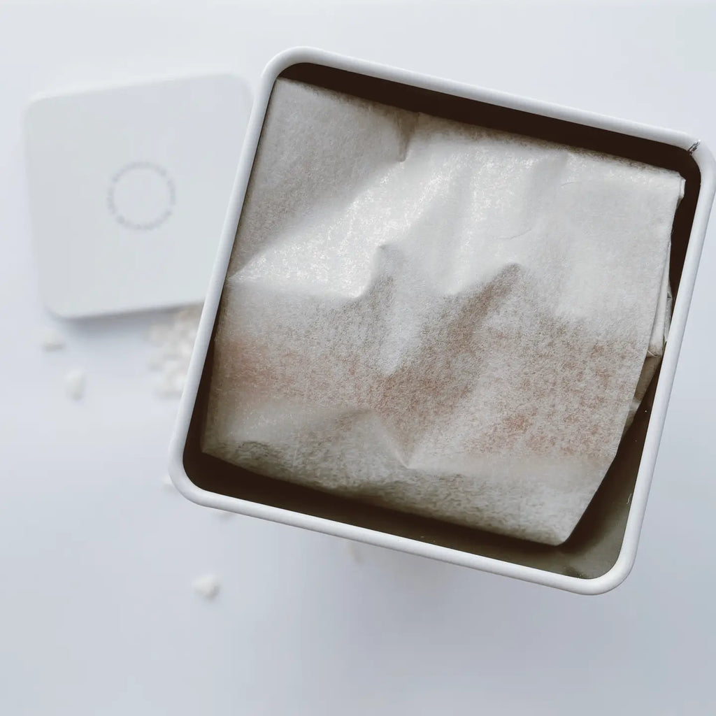 A white, square container of Mother Mother Magnesium Bath Soak with its lid removed reveals a paper-wrapped item inside. Some small white granules, likely from the magnesium soak, are scattered near the lid.