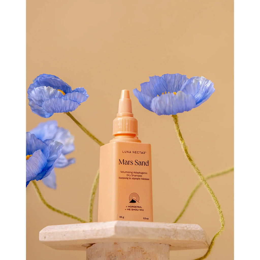 A bottle of luna nectar mars sand hair serum displayed on a pedestal with blue poppy flowers in the background.