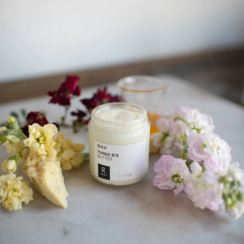 Jar of three b's butter skincare product surrounded by flowers on a marble surface.