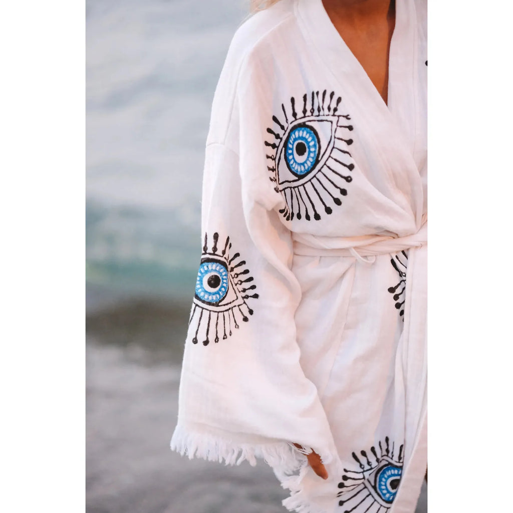 Person wearing a white robe with blue eye designs standing by the water.
