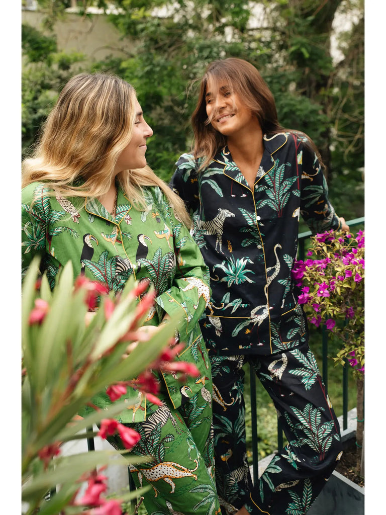 Two people stand on a balcony wearing classic and elegant patterned pajamas. One person is dressed in green, while the other is wearing Germaine des Prés Camille Black Jungle Print Pajamas. Their 100% cotton pajamas perfectly complement the lush potted plants and flowers surrounding them, with greenery stretching into the background.