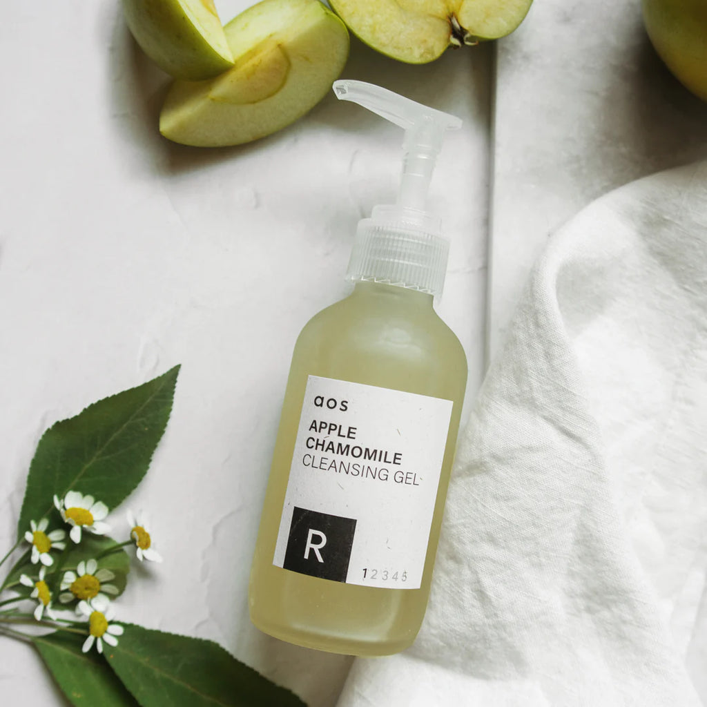 Bottle of apple chamomile cleansing gel surrounded by fresh apple slices and chamomile flowers on a light background.